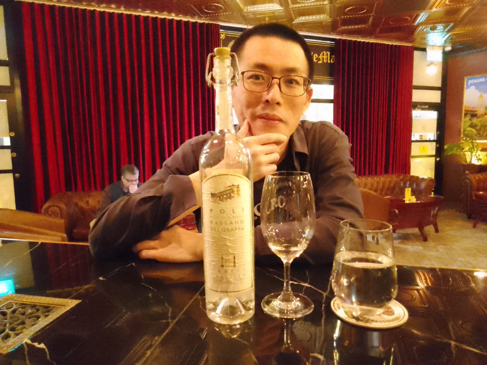photo of Tony at a bar that sells an Italian alcoholic beverage known as grappa. 
                 A young man smiles at the camera, with a backdrop that looks opulent.